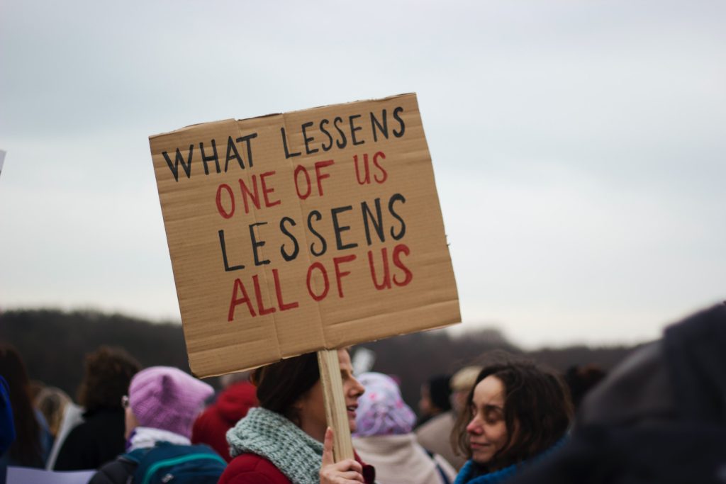 Une manifestante porte une pancarte avec le texte "what lessens one of us lessens all of us", en français "ce qui diminue l'un·e entre nous, diminue tou·te·s"